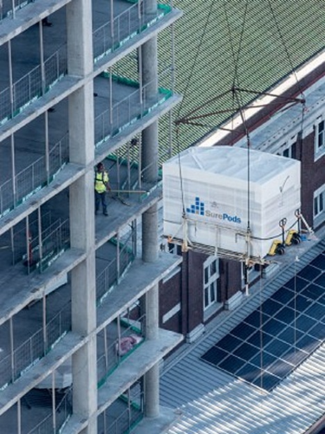 A crane hoists a SurePods bathroom unit into place on a high-rise construction site, showcasing modular building efficiency.