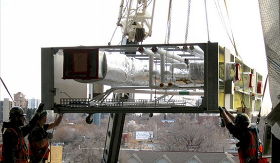 Construction workers guiding a prefabricated MEP rack into place with a crane at a construction site.